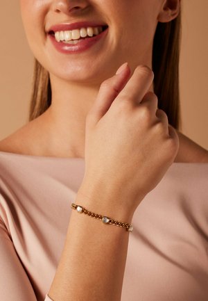 Woman smiling, wearing light pink top, showing gold chain bracelet with three white heart-shaped charms on wrist.