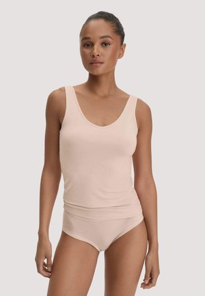 Woman wearing a light beige sleeveless tank top and matching underwear standing against a plain light background.