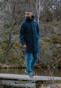 Cappotto lungo blu navy con cappuccio, dotato di chiusura con zip e polsini elastici, indossato sopra jeans blu chiaro e sneakers grigie, su un molo di legno.