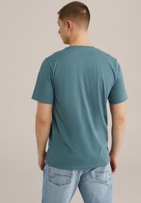 Rövid ujjú, türkiz színű pamut póló, kerek nyakkivágással, hátulról bemutatva. Világoskék farmernadrággal párosítva. Simító textúra, laza fazon.