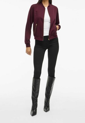Vrouw draagt een bordeauxkleurige jas met rits, witte top, zwarte skinny broek en zwarte leren kniehoge laarzen, staand tegen een witte achtergrond.