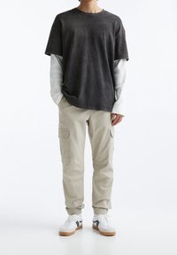 Dark gray short-sleeve shirt layered over a white long-sleeve shirt, paired with light beige cargo pants and white sneakers with black accents.