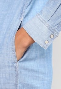 Světle modrá denimová košile s dlouhými rukávy, má knoflíkový manžet a boční kapsu. Tkanina má jemnou strukturu a neformální design.