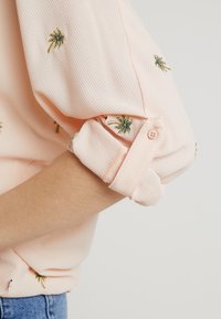 Jasnoróżowy, teksturowany rękaw zrolowany i zapięty, ozdobiony małymi haftowanymi wzorami palm w zielonym i złotym kolorze.