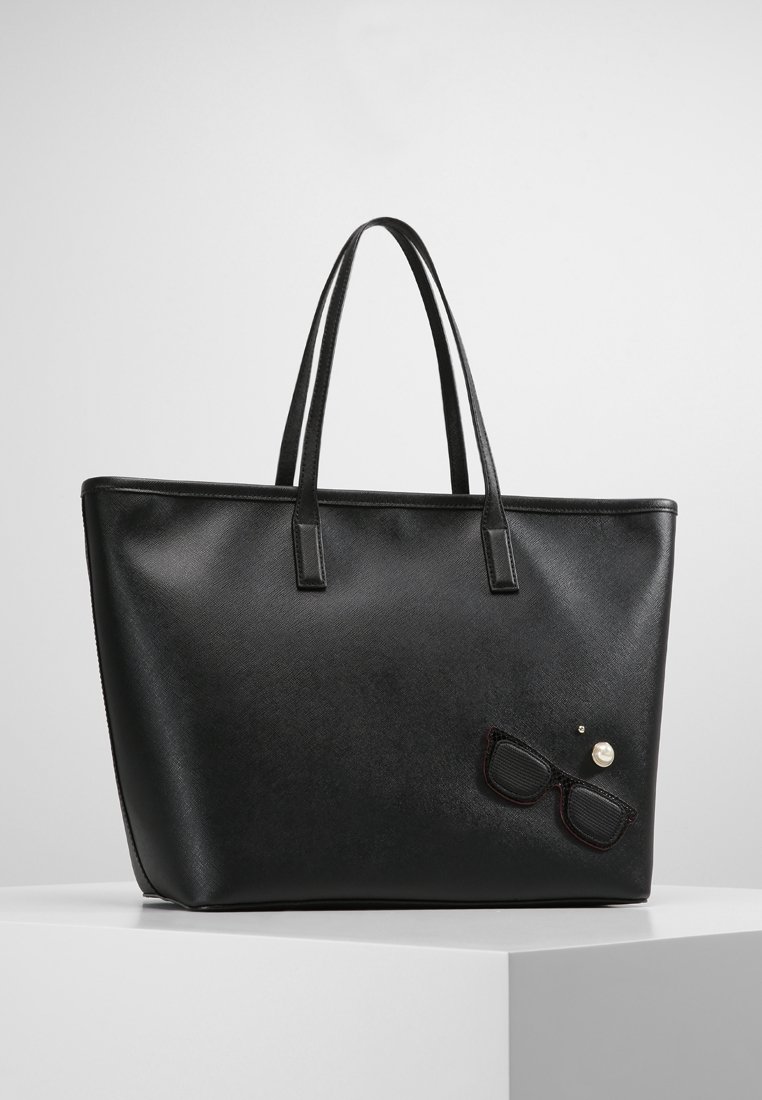 Sac cabas en cuir noir avec deux bandoulières et un petit patch décoratif de lunettes de soleil et une perle sur la partie avant inférieure droite.