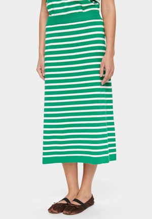 Falda midi de punto a rayas verdes y blancas con una cinturilla ajustada, textura suave y abertura lateral en el dobladillo. Combinada con zapatos planos marrones.