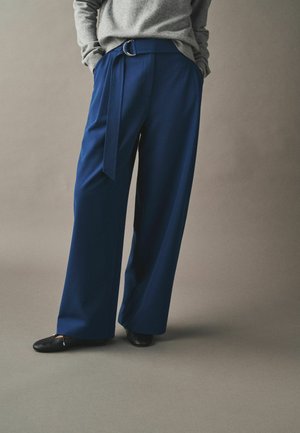 Person wearing loose blue trousers with a fabric belt and black flats, hands in pockets, standing against a plain grey background.