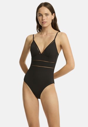 Kobieta z długimi brązowymi włosami, ubrana w czarny jednoczęściowy strój kąpielowy z cienkimi ramiączkami i siateczkowymi detalami na piersiach i talii, stoi na białym tle.