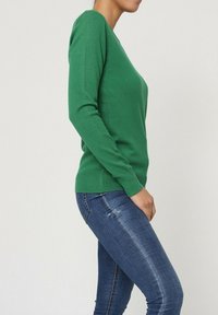 Sweater en tricot vert avec un col en V, des manches longues et une silhouette ajustée. Associé à un jean bleu, mettant en avant une texture lisse.
