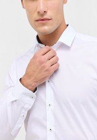 Camicia bianca con colletto accentato in nero. Presenta maniche lunghe, polsini con bottoni e un tessuto liscio e strutturato.