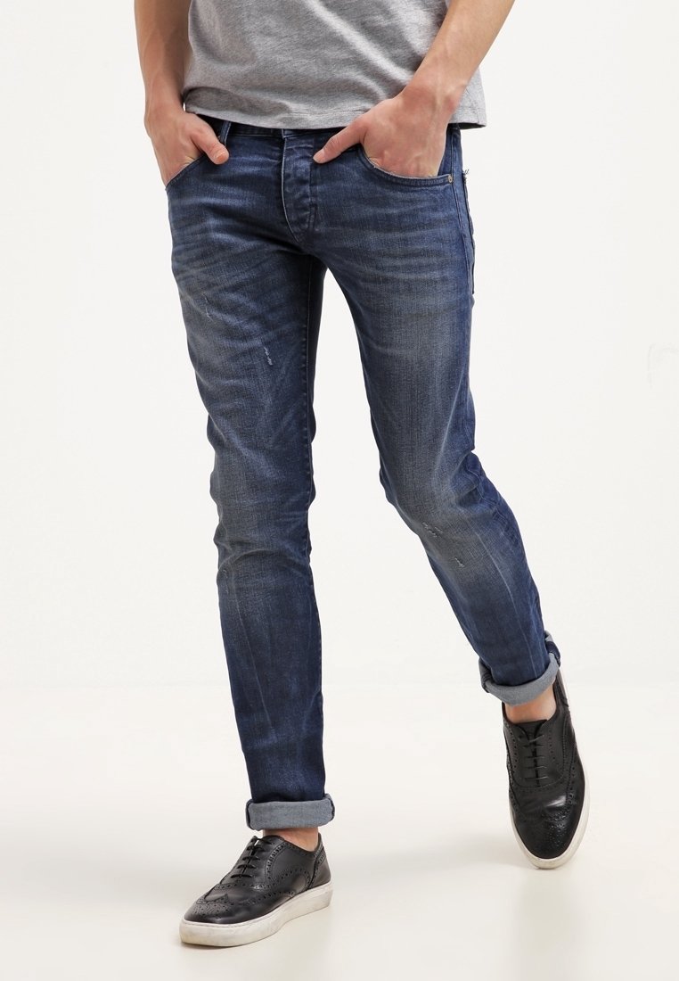 Man in opgerolde donkerblauwe jeans en zwarte leren brogue sneakers met witte zolen, handen in zijn zakken, staand op een witte vloer.