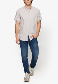 Helles graues kurzärmeliges Hemd mit Knopfleiste und Brusttasche, kombiniert mit blauen Jeans und beigen Sneakern, vor einem weißen Hintergrund stehend.