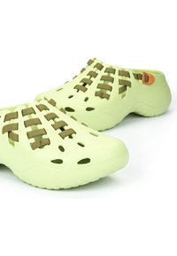 Zuecos de plástico verde claro con agujeros de ventilación recortados y correas tejidas en verde oliva. Presentan un pequeño acento naranja en el talón.