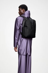 Czarny plecak o gładkiej teksturze, wyposażony w podwójne paski na ramiona i boczną kieszeń, noszony na błyszczącym fioletowym płaszczu przeciwdeszczowym i dopasowanych spodniach.