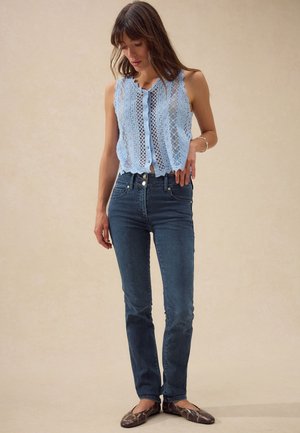 Žena nosi svetlomodrý čipkovaný top bez rukávov so zapínaním na gombíky, tmavomodré rifle s vysokým pásom a hnedé ploché topánky so vzorom zhadzej kože.