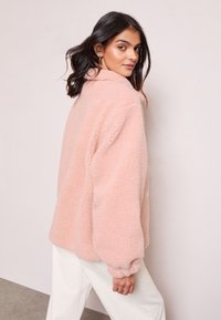 Růžová, nadýchaná fleecová bunda s vysokým límcem, elastickými manžetami a volným střihem, umístěná na neutrálním pozadí.