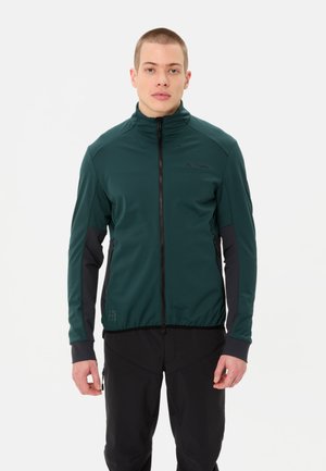 Junger Mann, der nach vorne blickt, trägt eine dunkelgrüne Zip-up-Jacke mit schwarzen Akzenten und schwarze Hosen vor einem schlichten weißen Hintergrund.