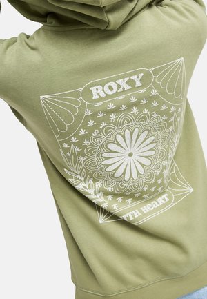 Osoba ubrana w oliwkowozieloną bluzę z dużym białym kwiatowym mandalą i napisem "ROXY" na plecach.