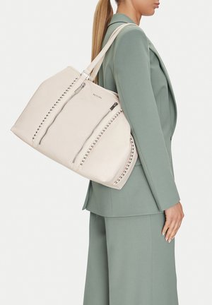 Mujer con traje verde salvia lleva un gran bolso tote color blanco roto con cremalleras verticales y detalles de tachuelas sobre el hombro.