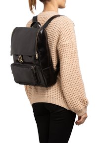 Vrouw in een beige gebreide trui en zwarte broek, met een zwarte leren rugzak met ritssluitingen op haar rug.