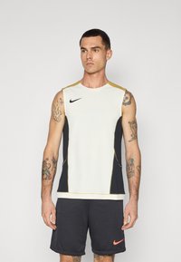 Moški brezrokavnik za šport: spredaj off-white, na straneh črn, z zlatimi poudarki. Ima mrežasto blago, logotip na prsih in kontrastne šive.