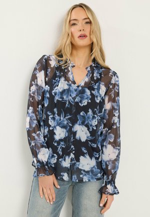 Blonde Frau trägt eine durchsichtige schwarze Bluse mit blauem und weißem Blumenmuster und hellblaue Jeans, steht vor einer schlichten weißen Wand.