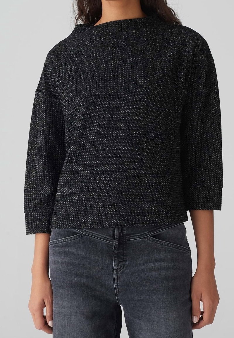 Maglia nera a tre quarti con tessuto testurizzato caratterizzato da sottili fili metallici. Rifinita con un collo a barca e una vestibilità rilassata.