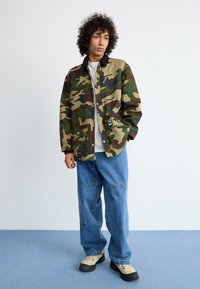 Καμουφλαρισμένο μπουφάν σε πράσινο, καφέ και μπεζ χρώμα, με μαύρο γιακά και κουμπιά τσίμπημα, συνδυασμένο με χαλαρές μπλε τζιν παντελόνες και μπεζ παπούτσια.