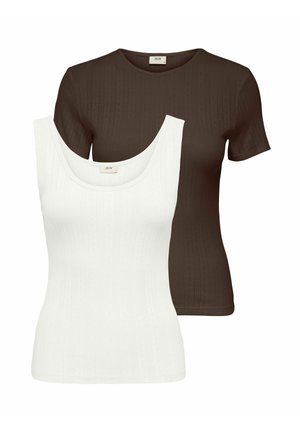 Femme souriante debout, les mains derrière le dos, portant un haut marron à manches courtes, un pantalon noir à jambes larges et des chaussures blanches pointues sur fond blanc.