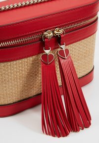 Czerwona skórzana torebka z plecionego słomy, wyposażona w zapięcie na suwak i dwa czerwone frędzle z sercowymi akcentami. Widoczne detale w odcieniu złota.