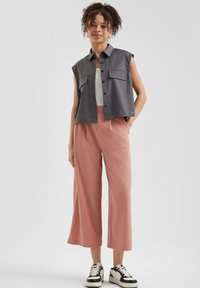 Sivá bezrukávová košeľa s dvoma náprsními vreckami, skombinovaná so svetloružovými širokými nohavicami. Čierno-biele tenisky dopĺňajú outfit.