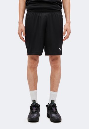 Osoba ubrana w czarne krótkie spodenki sportowe z logo Puma, białe skarpetki za kostkę i czarne buty sportowe, stojąca na jednolitym tle.