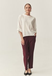 Vrouw met haar opgestoken in een losse witte blouse, bordeauxrode broek, gouden creolen en zwarte puntige flats, staand met één hand in de zak.