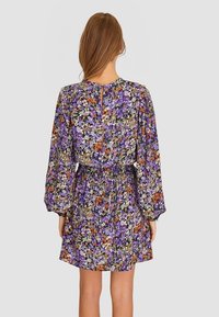 Vestido floral con un patrón en púrpura, blanco y naranja, que presenta mangas largas, cintura elástica y un detalle de escote en forma de llave en la parte posterior.