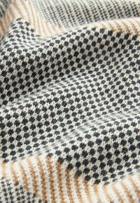 Tessuto di lana intrecciato con un motivo pied de poule nei colori nero, bianco e beige. Superficie testurizzata con variazioni di colore e motivo ben visibili.