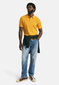 Gelbes Polohemd, blaue Jeans und braune Sneaker mit sandfarbenen Akzenten; grüner Sweatshirt um die Taille gebunden. Lässiges Outfit mit klassischer Passform.