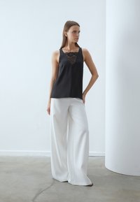 Top negro sin mangas con un inserto de encaje, combinado con pantalones anchos blancos, sobre un fondo minimalista. Textura suave y diseño simple.