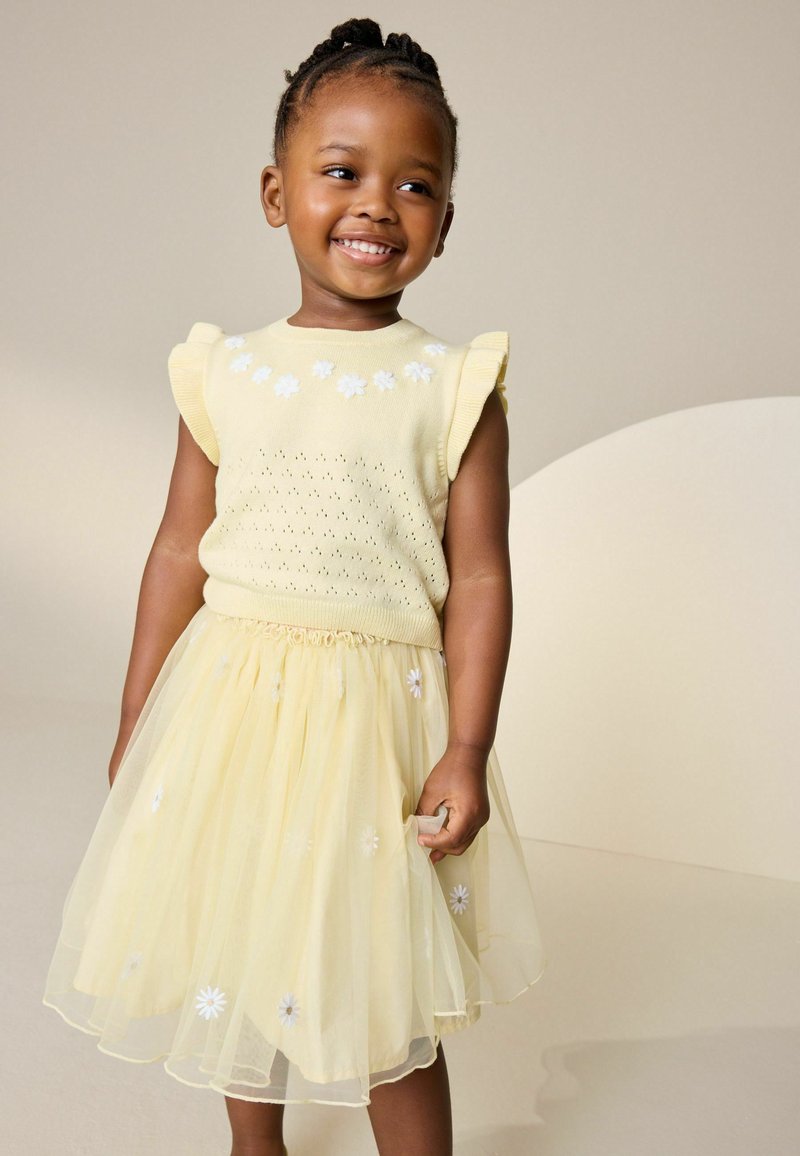 Uśmiechnięta młoda dziewczyna z zaplecionymi włosami, ubrana w żółtą bezrękawnik i pasującą tiulową spódnicę ozdobioną białymi kwiatami.