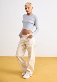 Hellgrau gestreifter Crop-Top mit langen Ärmeln, kombiniert mit cremefarbenen weiten Hosen, getragen mit weißen Sneakern. Einfaches Design mit minimalen Akzenten.