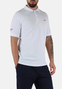 Polo shirt bianca con maniche corte, colletto e accenti blu chiaro a contrasto. Presenta un logo sul petto e testo rosso sulla manica.