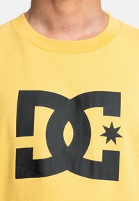 Žlté bavlnené tričko s veľkým čiernym grafickým logom so detailom hviezdy, okrúhlym výstrihom a textúrovým materiálom.