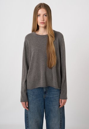 Junge Frau mit langen, glatten Haaren, die einen lockeren grauen Pullover und blaue Jeans trägt und vor einem schlichten hellen Hintergrund steht.