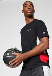 Camiseta negra de manga corta con paneles laterales rojos y blancos, que presenta un pequeño logo bordado en el pecho, combinada con pantalones cortos negros y sosteniendo un balón de baloncesto.