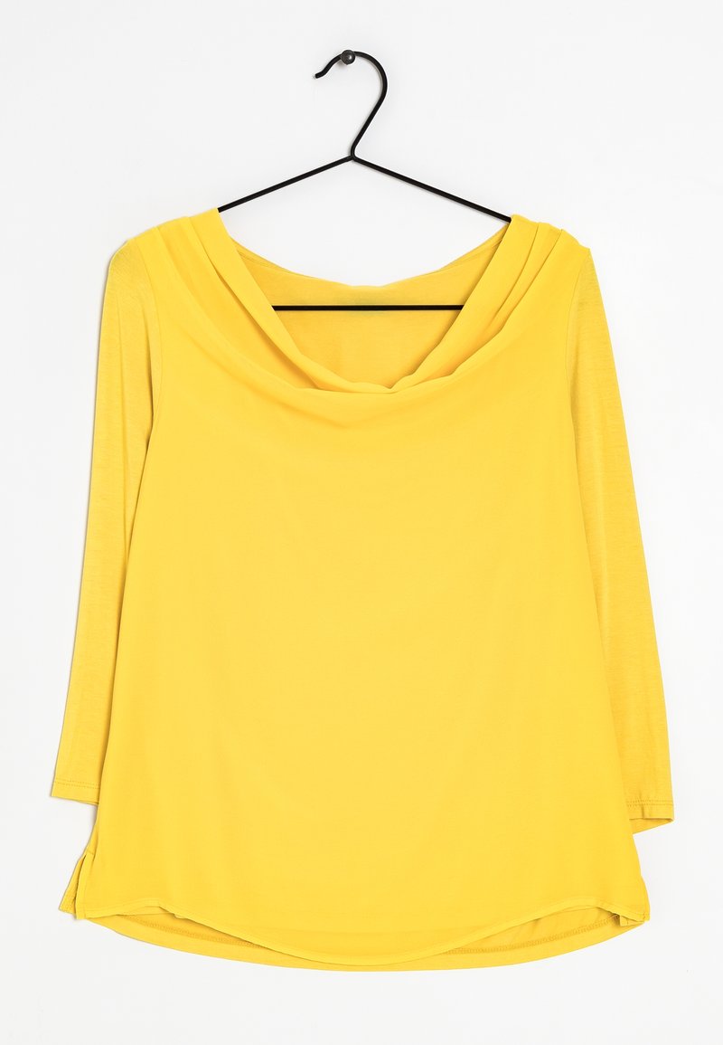 Jasnożółta bluzka z długim rękawem z drapowanym dekoltem w kształcie cowl, wisząca na prostym czarnym metalowym wieszaku na białym tle.