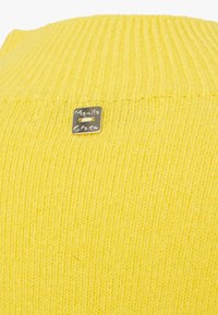 Maglione giallo a maglia con texture a coste, dotato di un piccolo logo metallico cucito sul colletto.