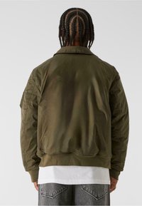 Olivgrön bomberjacka tillverkad av ett slätt material, med ribbat fåll och krage, subtil lyster och sidfickor.