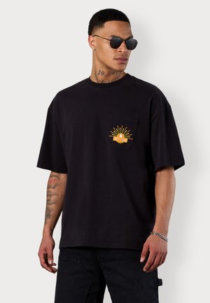 Schwarzes Baumwoll-T-Shirt mit lockerer Passform, ausgestattet mit einer Brusttasche und einem bunten Sonnenmotiv. Getragen mit schwarzen Jeans und Sonnenbrille.