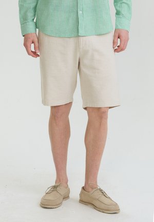 Mand iført beige knælange shorts, beige sko og en grøn-hvid stribet skjorte med knapper står på en hvid gulv.