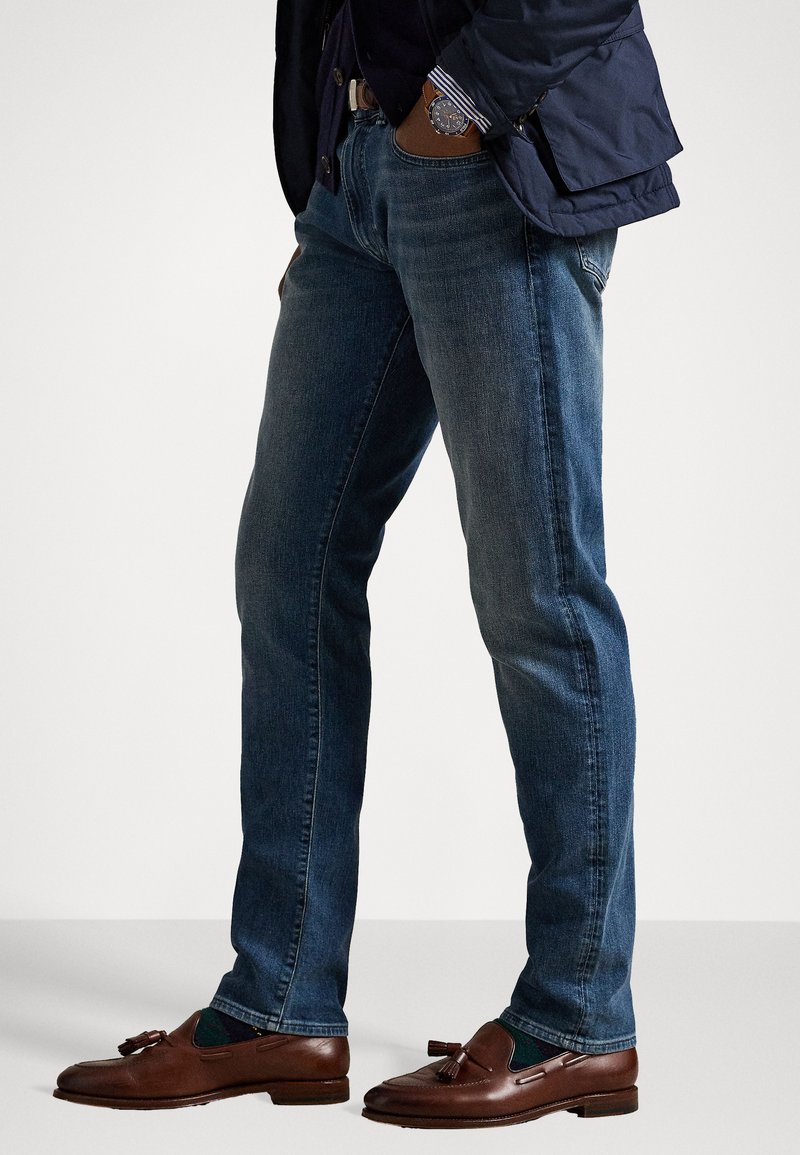 Dark blue denim jeans with a straight cut, featuring a classic five-pocket design. Paired with brown leather loafers and a navy jacket.