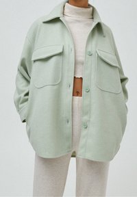 Shacket oversize verde chiaro realizzato in tessuto morbido, con due tasche sul petto, chiusura frontale con bottoni e colletto appuntito.